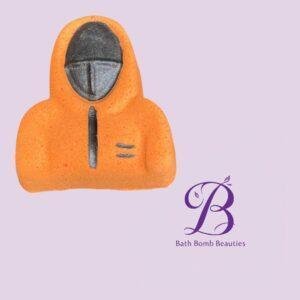 Orange hooded bath bomb resembling a person with a dark face mask on a lavender background; Bath Bomb Beauties logo in the corner.