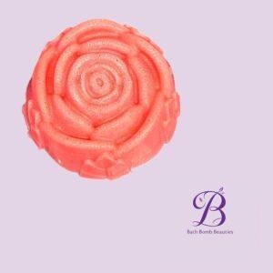 Close up of a pink rose shaped bath bomb with subtle glitter on a lavender background bath bomb beauties logo in purple at bottom right