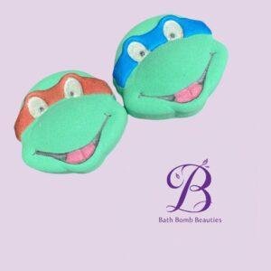 Two green frog-shaped bath bombs with red and blue heads and smiling faces sit on a lavender background, with a purple Bath Bomb Beauties logo in the corner.