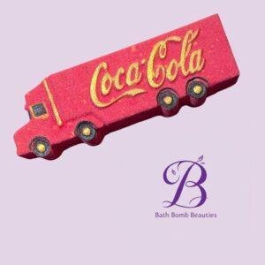 Red Coca-Cola–style truck-shaped bath bomb on a pale lavender background, with Bath Bomb Beauties branding visible.