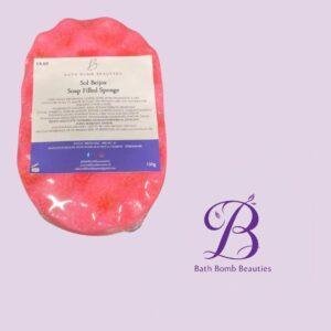 Pink bath bomb sponge with label reading 'Sol Beijos Soap Filled Sponge' placed next to a purple 'Bath Bomb Beauties' logo on a lavender background, product shot for branding and sale.