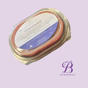Close-up of a glass jar with a metal lid containing a red/orange bath bomb, set against a light purple background, with Bath Bomb Beauties branding visible on the label and backdrop.