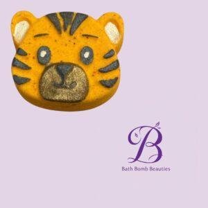 Tiger-shaped yellow bath bomb with black stripes and gold nose on a lavender background, beside the Bath Bomb Beauties logo, branding visible.