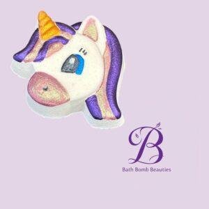 Unicorn shaped bath bomb with purple mane pink snout and a gold horn on a light purple background bath bomb beauties logo visible