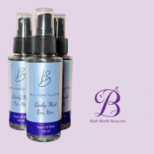 Three spray bottles with black caps and light blue labels reading bath bomb beauties body mist for her touch of pink  ml set against a lavender background with a purple b logo