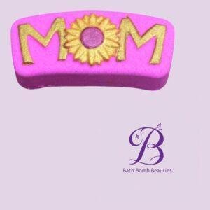 Pink curved bath bomb with the letters M-O-M, a gold sunflower as the O, and the Bath Bomb Beauties logo in purple at the bottom right.