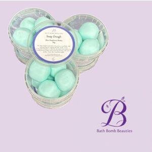 Three clear containers filled with turquoise bath bombs arranged on a pastel lavender background with the bath bomb beauties logo visible