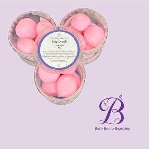 Pink bath bombs labeled soap dough candy floss in clear round containers on a lavender background