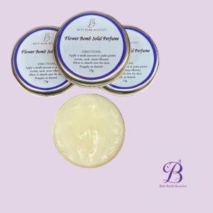 Three metal tins with blue labels reading 'Flower Bomb Solid Perfume' on a pale lavender background; Bath Bomb Beauties logo visible.