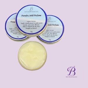 Three blue-labeled Paradox Solid Perfume tins with usage directions, on a lavender background; Bath Bomb Beauties logo bottom right.
