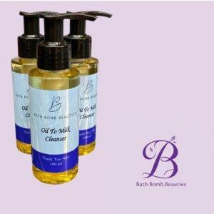 Three Bath Bomb Beauties oil-to-milk cleanser bottles with pump dispensers on a lavender background; the labels read 'Oil To Milk Cleanser'.