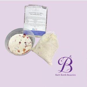 Bath bomb ingredients in a metal bowl with dried flower petals, a paper bag and a cloth pouch on a lavender background with a decorative logo.