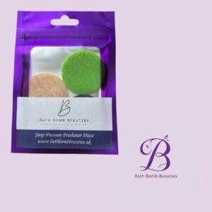 Purple Bath Bomb Beauties package showing two round discs (green and peach) visible through a window on a lavender background; branding and logo present on the packaging.