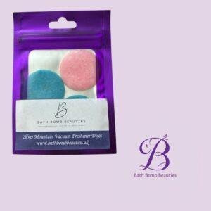 Purple pouch labeled Bath Bomb Beauties with blue and pink vacuum freshener discs visible; front label shows logo and website, purple background.