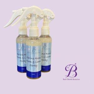 Three white spray bottles with blue labels reading bath bomb beauties on a lavender background logo bottom right