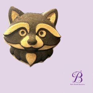 Close up of a black and gold raccoon themed cookie on a lavender background with bath bomb beauties logo in the corner